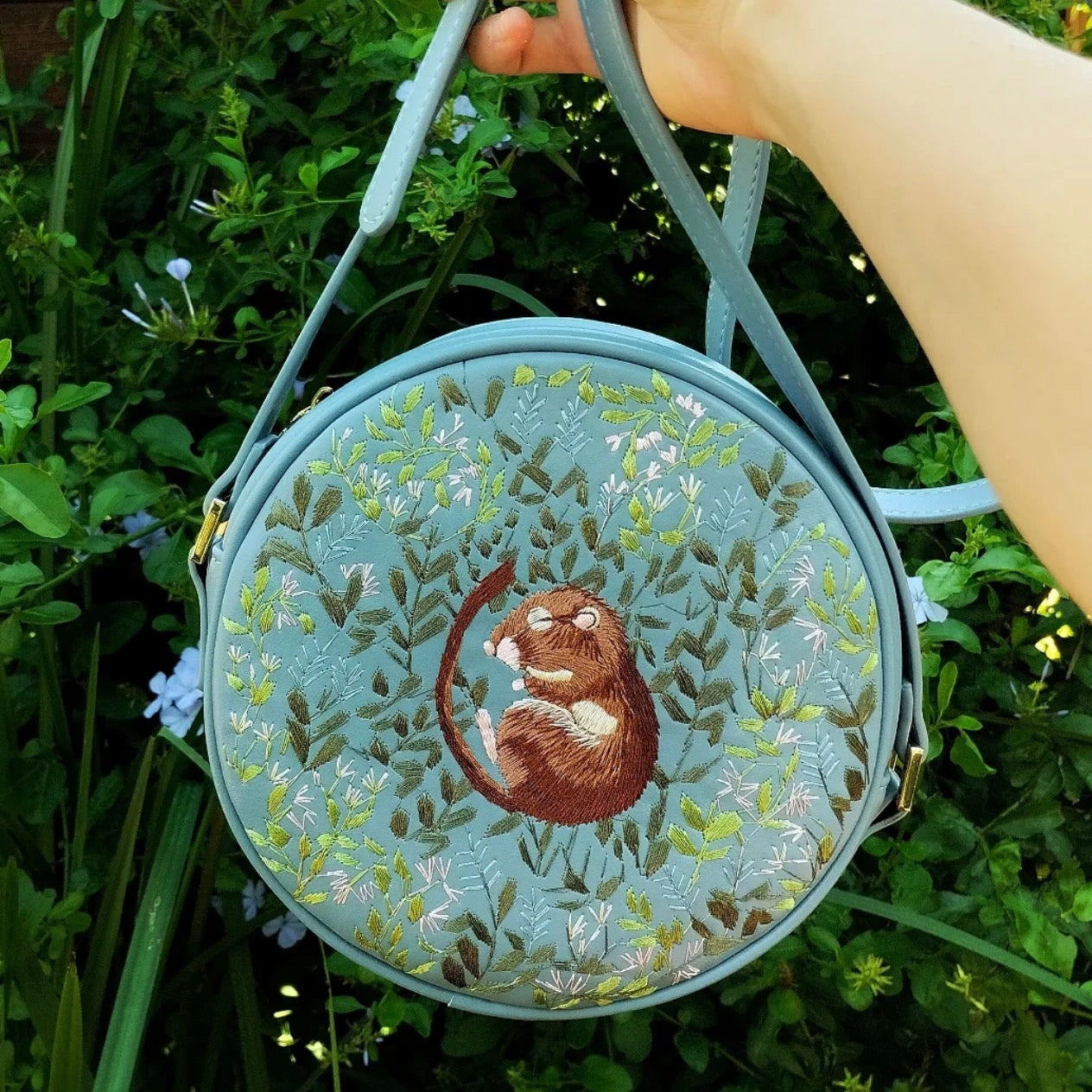 Round embroidered bag with a design of a mouse and leaves held by a hand against a green background.
