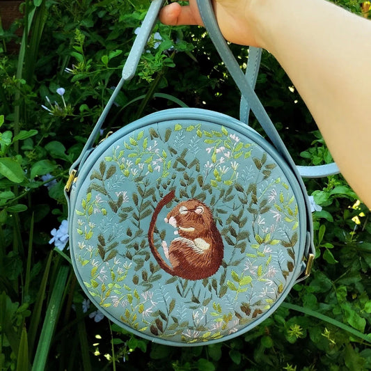 Round embroidered bag with a design of a mouse and leaves held by a hand against a green background.